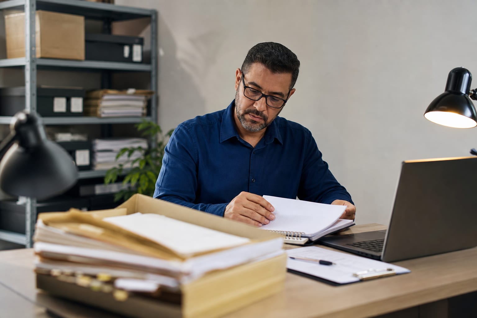 Contador consultando anotações ao lado do computador com pastas de arquivo abertas sobre a mesa do escritório